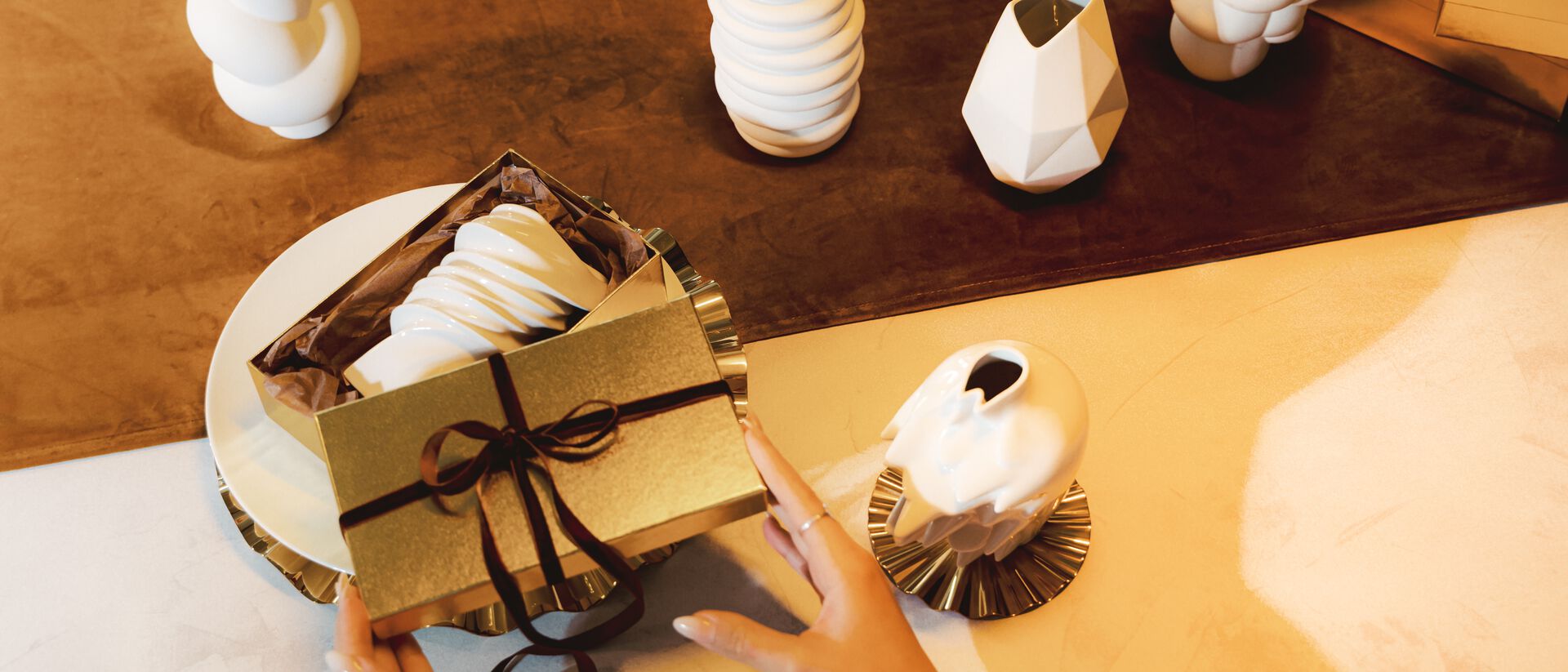 Hände öffnen eine goldene Geschenkbox mit modernem weißen Porzellan, umgeben von dekorativen Designer-Vasen von Rosenthal auf einer festlich gedeckten Tafel.
