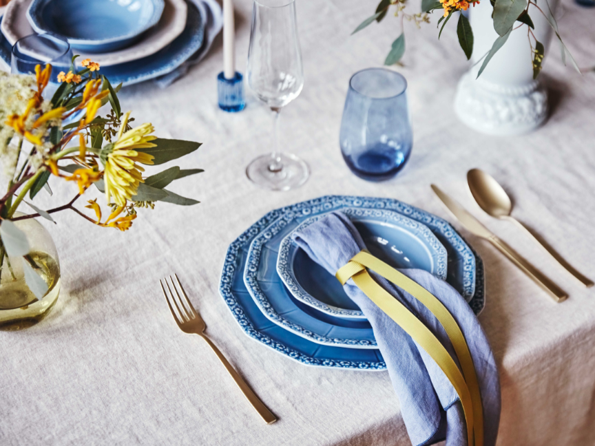 Stilvoll gedeckter Tisch mit blauem Rosenthal Maria Porzellan, goldfarbenem Besteck, Stoffserviette mit gelbem Band und frischen Blumen in zarten Vasen