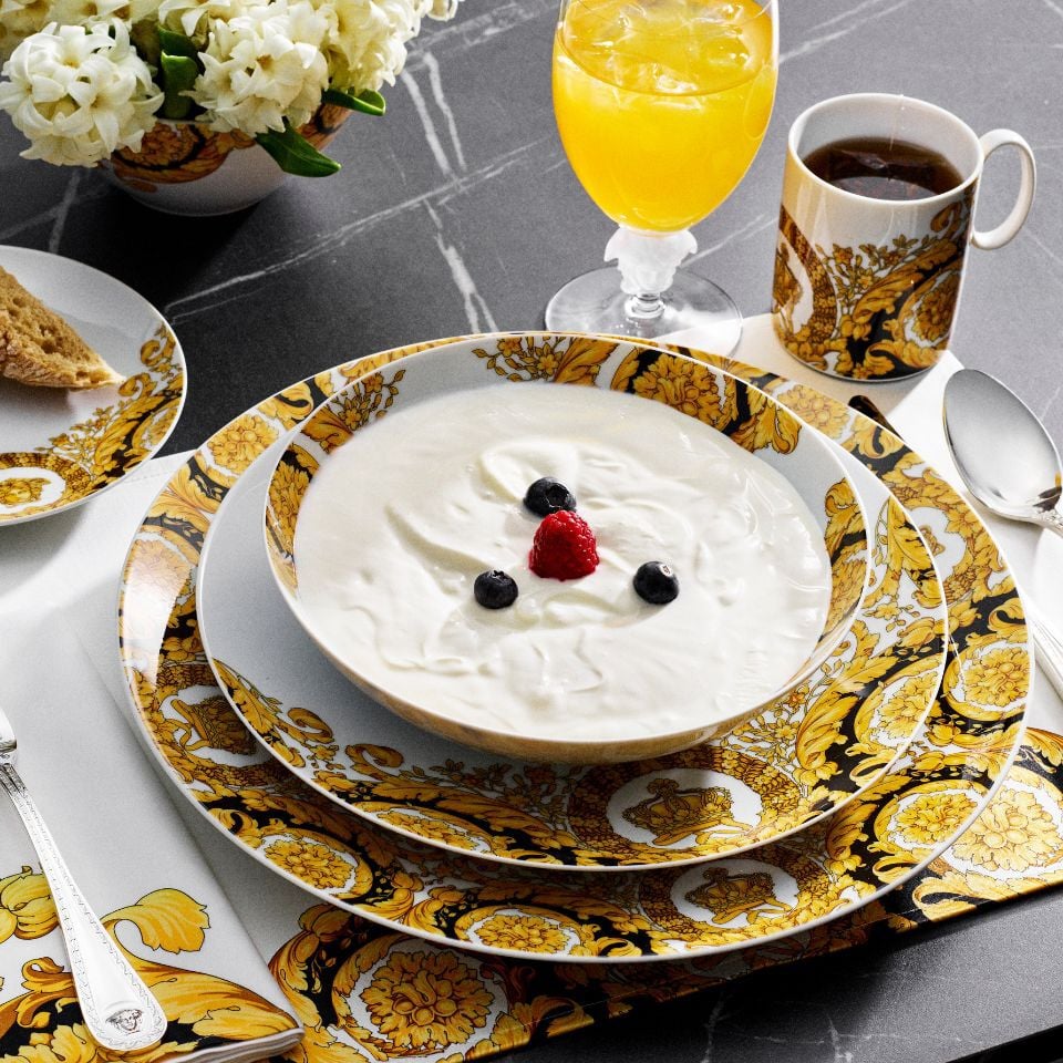 Barocco '92 porcelain breakfast set by Versace in yellow and gold baroque patterns, served with yogurt, berries, orange juice, and black coffee on a black marble table.