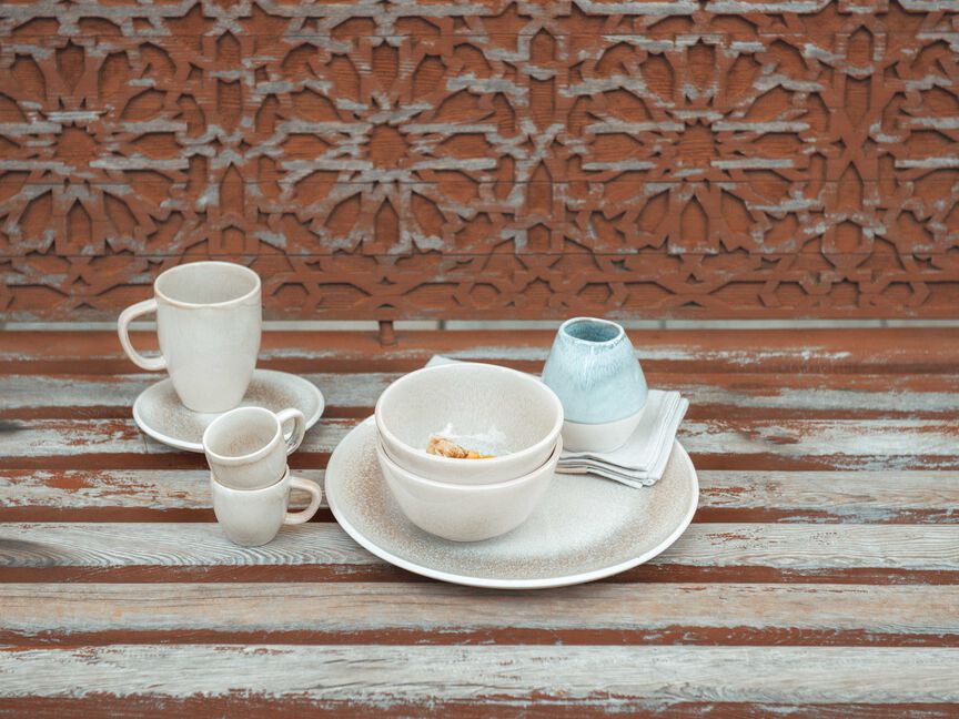Stoviglie in gres di Rosenthal: tazze, ciotole e piatti nei toni del crema e pastello su una panca rustica in legno davanti a una parete decorativa in metallo.