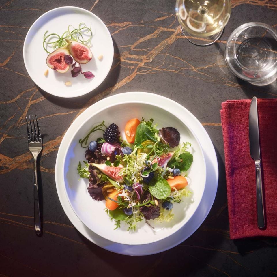 A white porcelain plate filled with a colorful mixed salad of greens, fruits, and figs, served on a dark stone-pattern table with a small side plate of sliced figs and a glass of white wine.