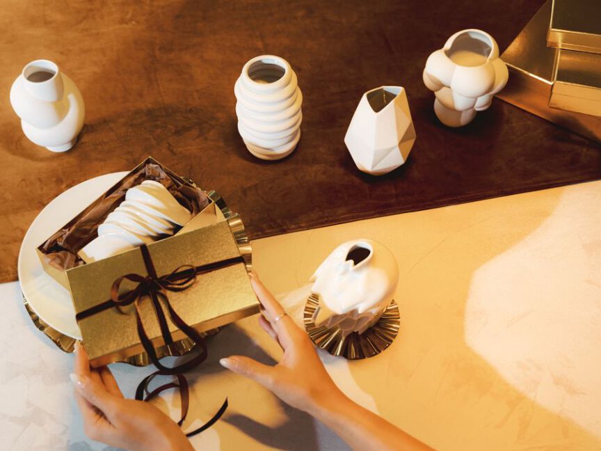 Ein goldenes Geschenk wird geöffnet, in dem eine weiße Midivase liegt. Auf dem Tisch stehen weitere Vasen von Rosenthal.