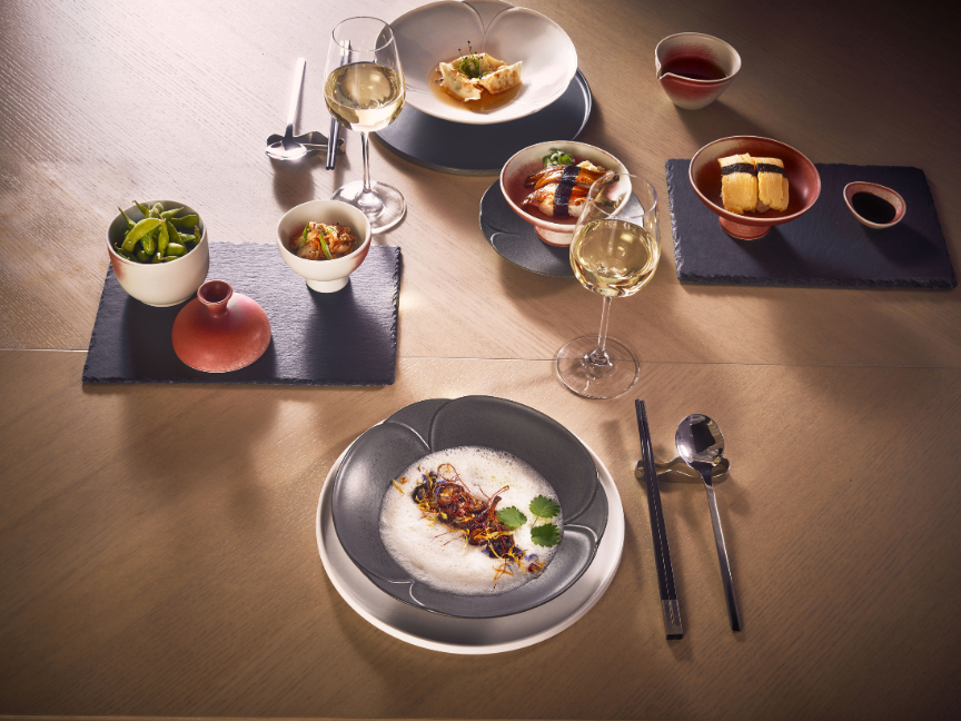 Gedeckter Tisch mit grauem Geschirr und Schiefer-Platzsets, auf denen asiatisch inspirierte Gerichte in Schalen und auf Tellern angerichtet sind.