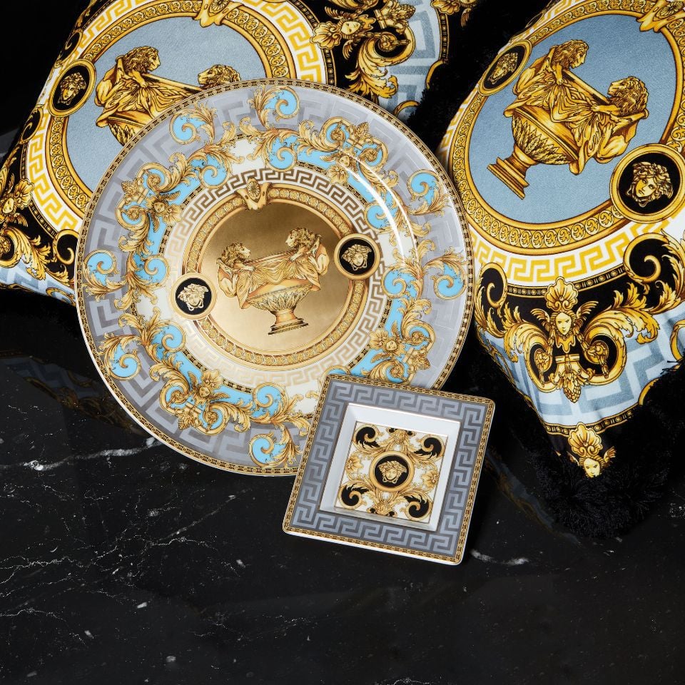 Prestige Gala plate and cushion set by Versace, showcasing classical vase motifs and blue-gold baroque designs with geometric borders on a black marble background.