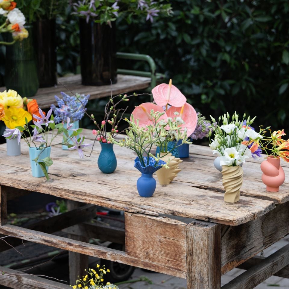 A rustic wooden table with small colorful vases holding wild and exotic flowers, set in a garden.