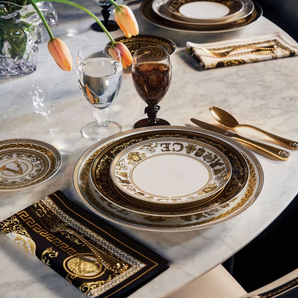 Versace Virtus Gala porcelain dinnerware with rich golden baroque and mythological motifs, set elegantly with gold flatware and tulips on a white marble table.
