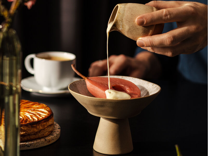Eine Hand hält einen Gießer, aus dem Vanillesoße in eine Schale mit Fuß von Rosenthal Sonetto Stoneware auf eine Birne fließt.