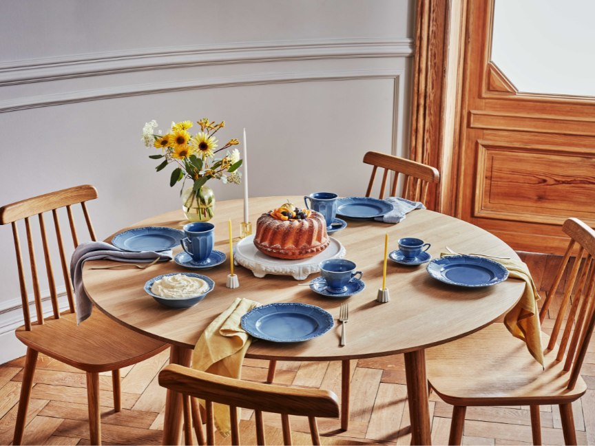 Einladend gedeckter Holztisch mit blauem Rosenthal-Geschirr, Gugelhupf auf Tortenplatte, Sonnenblumenstrauß und vier Gedecken für Kaffee und Kuchen