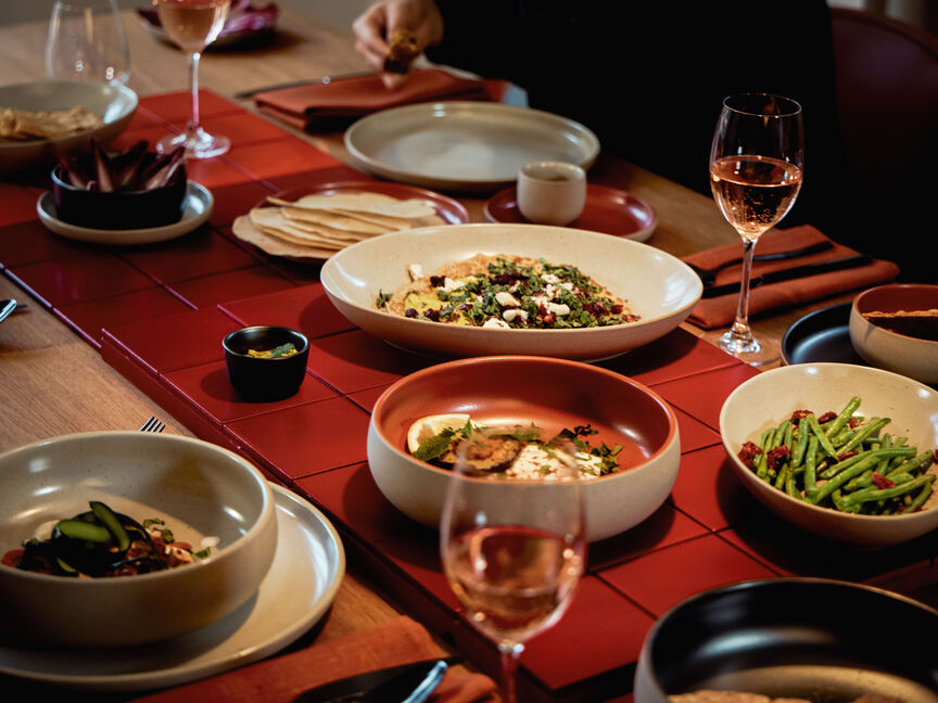 Tavola apparecchiata con stoviglie in gres Joyn di Rosenthal: diverse ciotole e piatti con vari piatti su un runner rosso, accompagnati da bicchieri e posate.