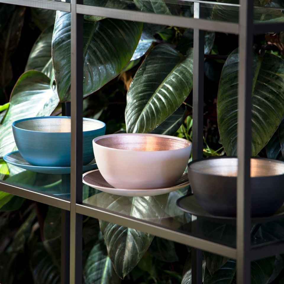 Three glass bowls in teal, blush and charcoal with matching plates, displayed on glass shelves.