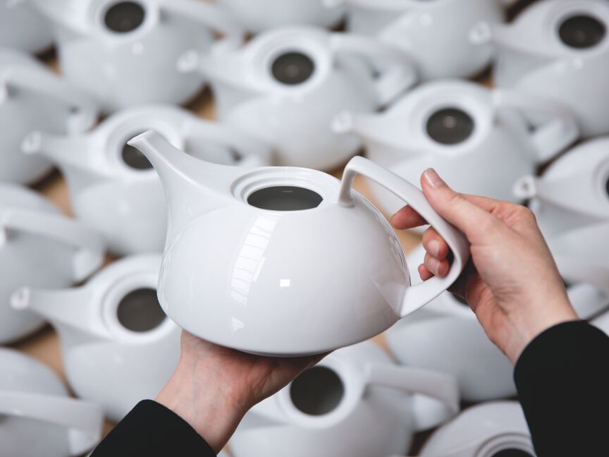 Eine Hand h&auml;lt eine fertige wei&szlig;e Teekanne, w&auml;hrend im Hintergrund zahlreiche identische Kannen f&uuml;r die Qualit&auml;tskontrolle bereitstehen.
