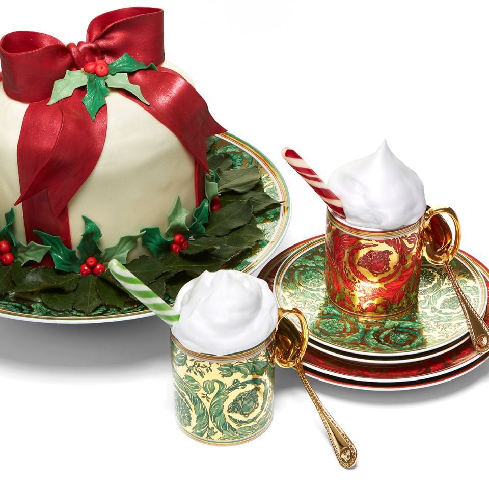 Festive Versace Medusa Garland collection with red and green baroque-patterned mugs and plates, featuring gold handles and paired with a decorative Christmas cake topped with a large red bow.