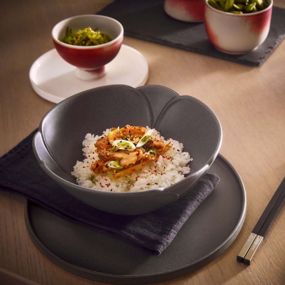 A grey ceramic bowl filled with rice and stir-fried vegetables, placed on a matching grey plate with dark chopsticks beside it. In the background, smaller red and white bowls contain side dishes.