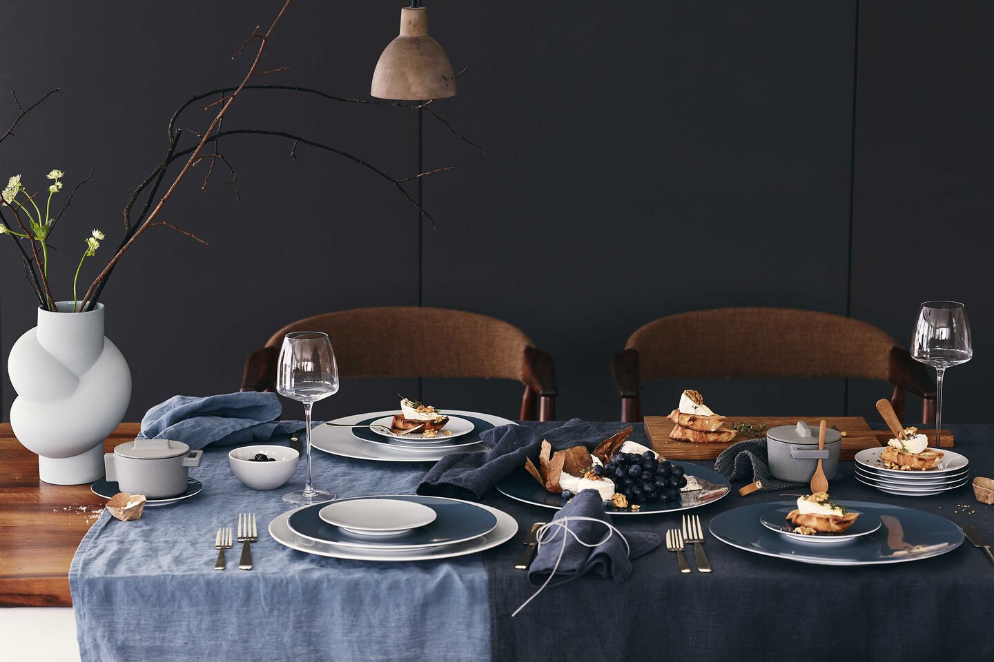 Rosenthal Dining Collection TAC Sensual Wooden table set with dark blue linen tablecloths and napkins with plates from the TAC Sensual Dining Collection, glasses, cutlery, snacks served and a white Node vase.