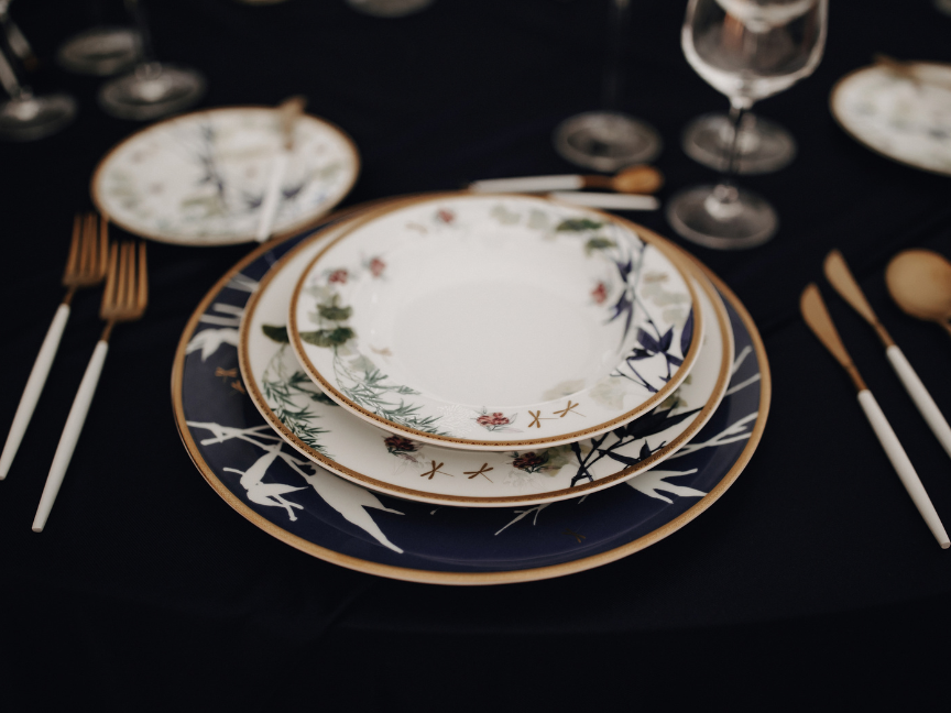 Festlich gedeckter Tisch mit Porzellantellern mit Blumendekor und Goldrand auf dunkelblauem Platzteller, dazu goldenes Besteck mit wei&szlig;en Griffen und Weingl&auml;ser.
