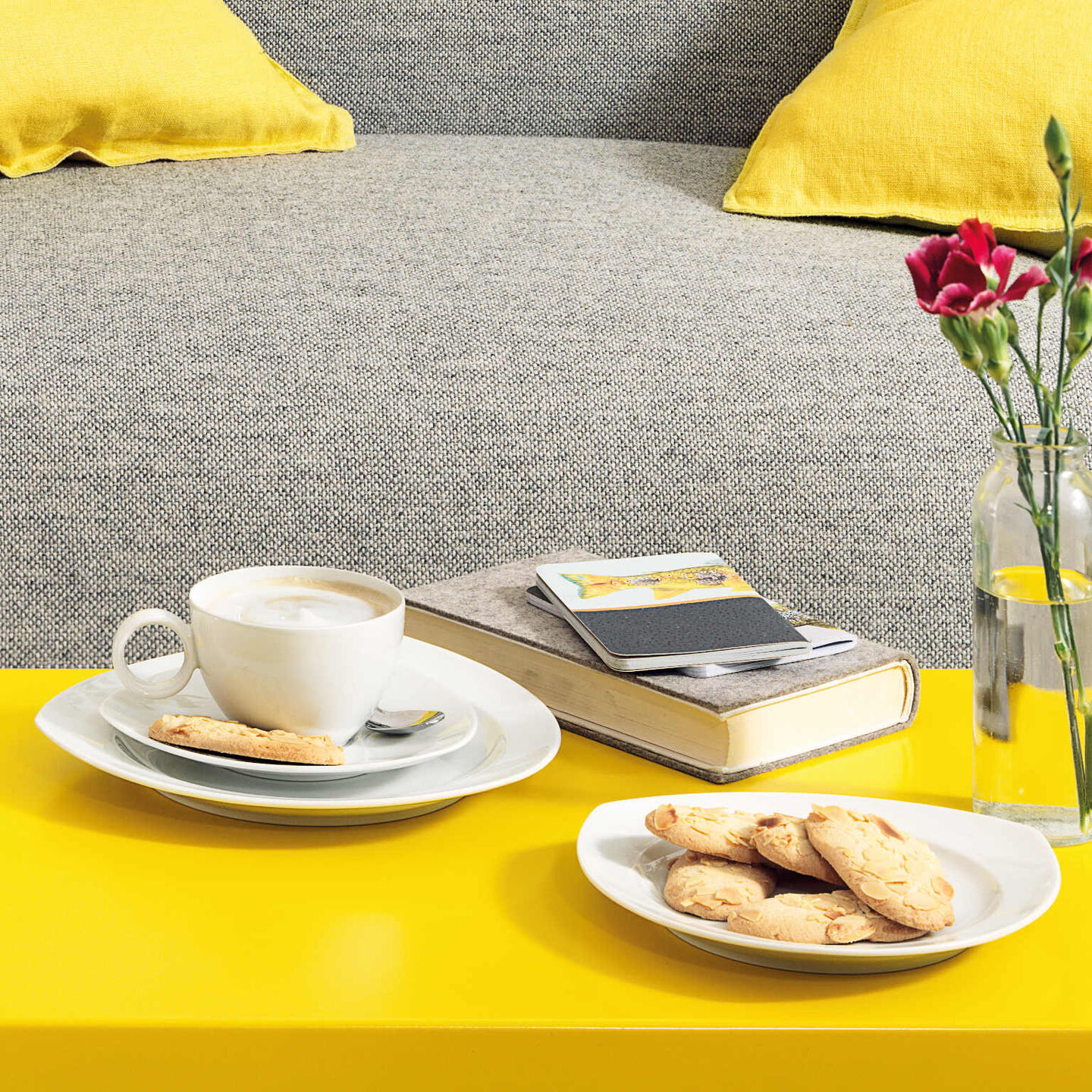 Thomas Vario cup filled with capuccino, plates with cookies on yellow couch table in front of a grey couch