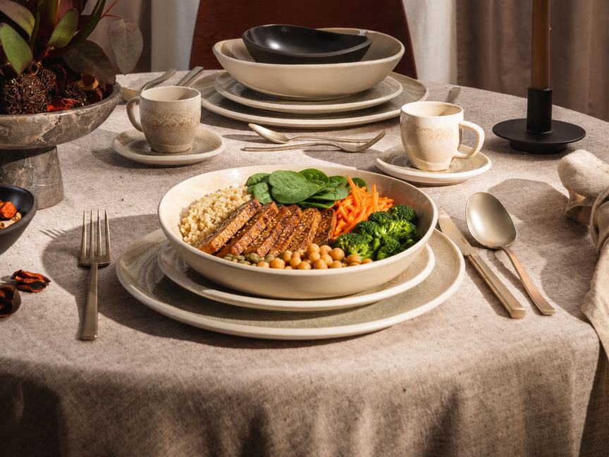 A laid table with cream-coloured crockery and a bowl of rice, fried tempeh, carrots, broccoli and spinach, with cutlery and cups beside it.