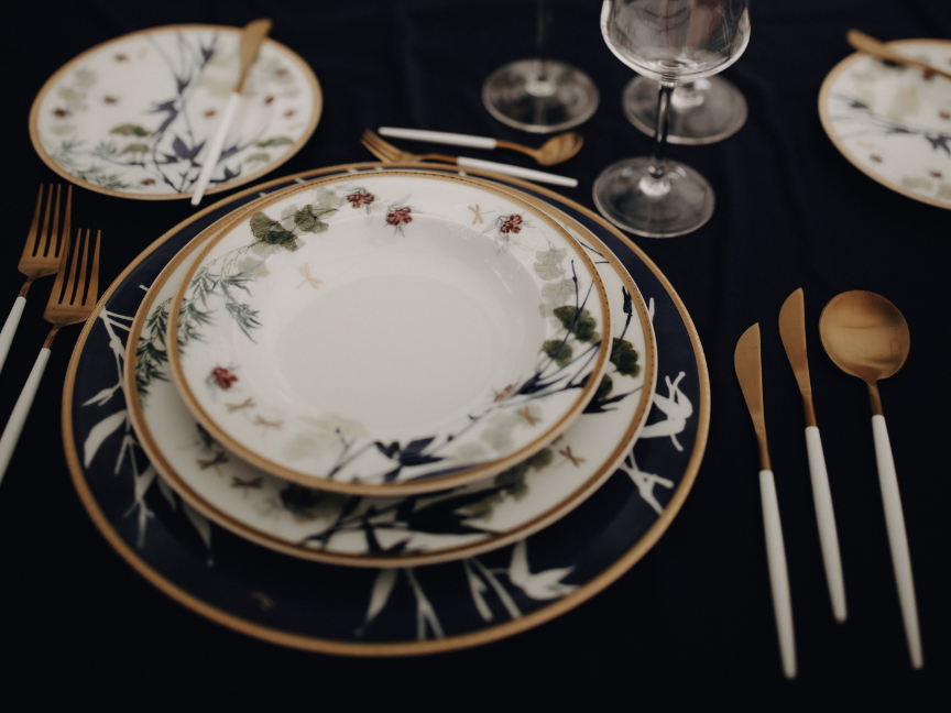 Stilvoll gedeckter Tisch mit floral verziertem Rosenthal-Porzellan, goldgerahmten Tellern, modernen Gläsern und goldfarbenem Besteck auf dunkler Tischdecke