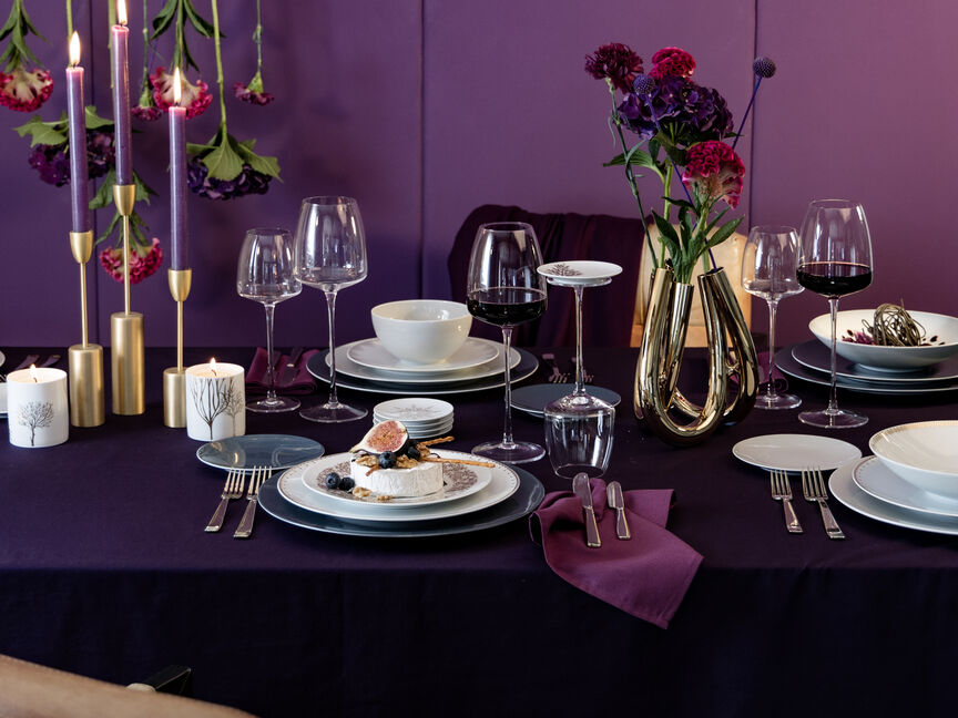 Elegant gedeckter Weihnachtstisch in Violett- und Goldtönen mit Kerzen, Blumenarrangement, edlem Porzellan und Weingläsern.