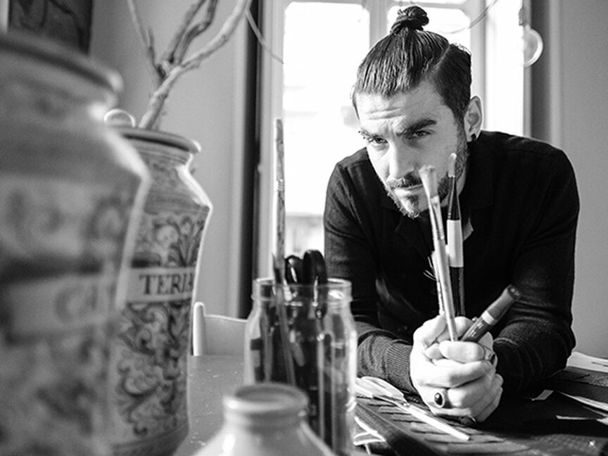 Black-and-white photograph of Designer Gianni Cinti in a studio, thoughtfully holding paintbrushes –