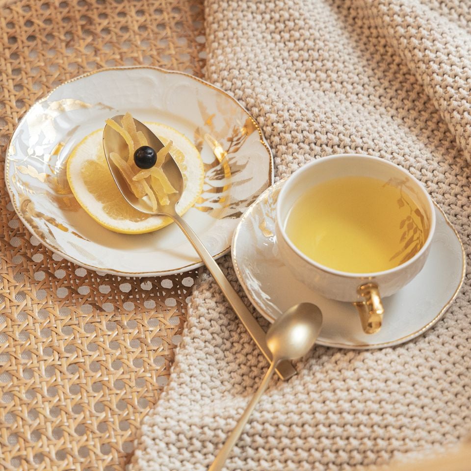 A cup of light-colored tea and a side plate with a lemon slice and candied ginger, served on elegant white porcelain with gold accents, set on a woven beige surface next to a knitted blanket.