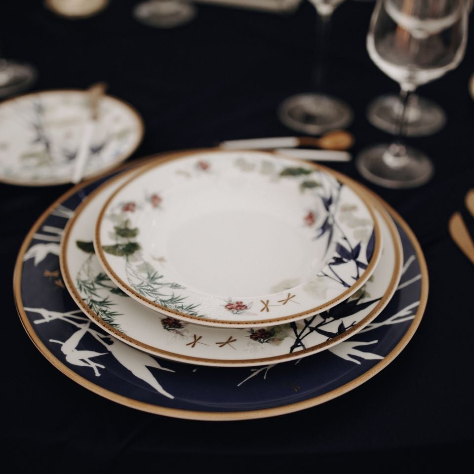 Layered porcelain plates with detailed floral and branch patterns in white, green, and navy tones, styled on a dark tablecloth with glassware and golden cutlery.
