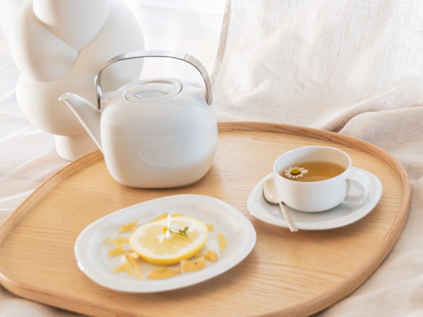 Wei&szlig;e Teekanne mit Metallgriff, eine Tasse Tee und ein Teller mit Zitronent&ouml;rtchen auf einem runden Holztablett vor hellem Stoffhintergrund.