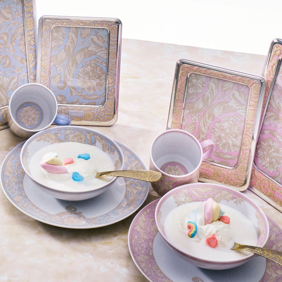 Soft pastel-toned Versace Kids porcelain breakfast set with pink and blue baroque designs, bowls of cereal with marshmallows, and matching frames on a marble surface.