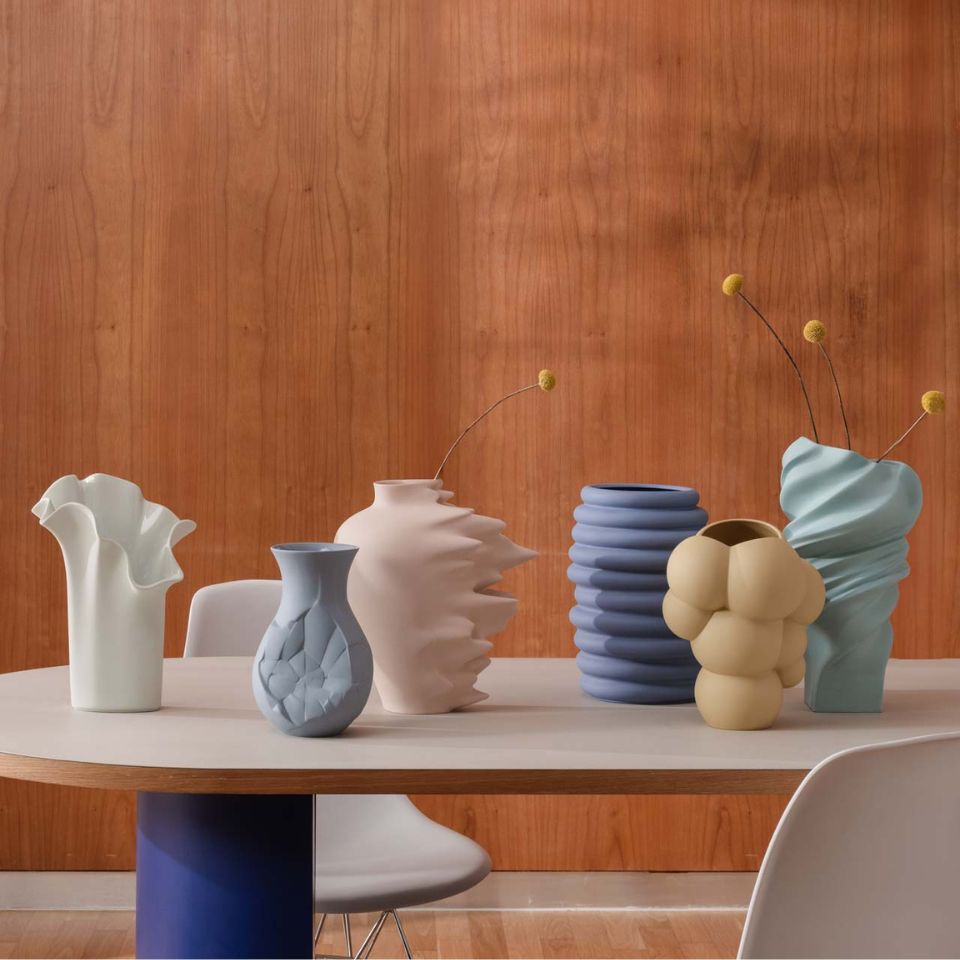 A row of sculptural matte vases in pale pastel colors arranged on a modern table in front of a wooden wall.