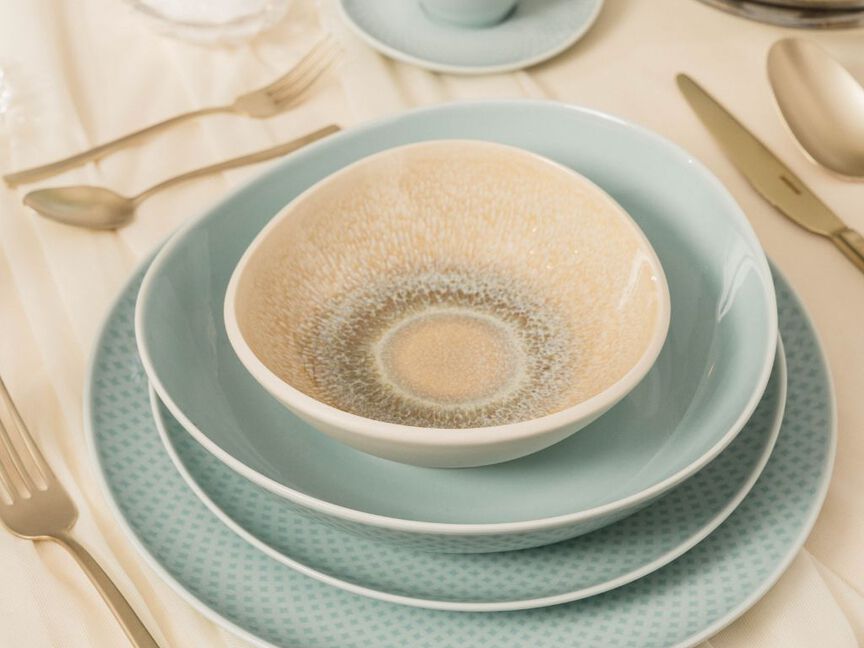 Stilvoll gedeckter Tisch mit mehrlagigem Rosenthal-Geschirr in sanften Blau- und Beigetönen, umgeben von elegantem Besteck auf cremefarbener Tischdecke.