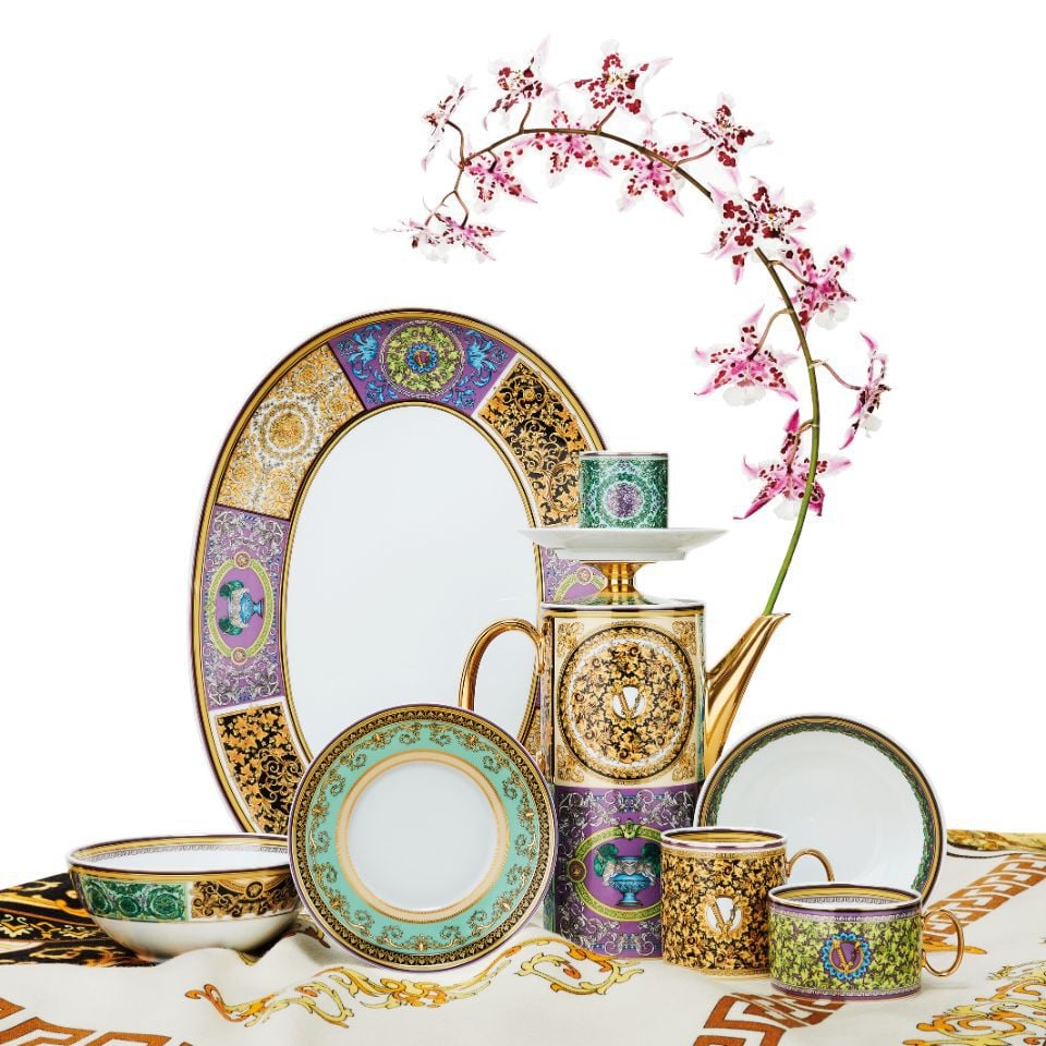Barocco Mosaic fine porcelain tableware set by Versace featuring a multicolored patchwork of intricate medallions, gold detailing, and regal motifs, elegantly arranged with a pink orchid flower.