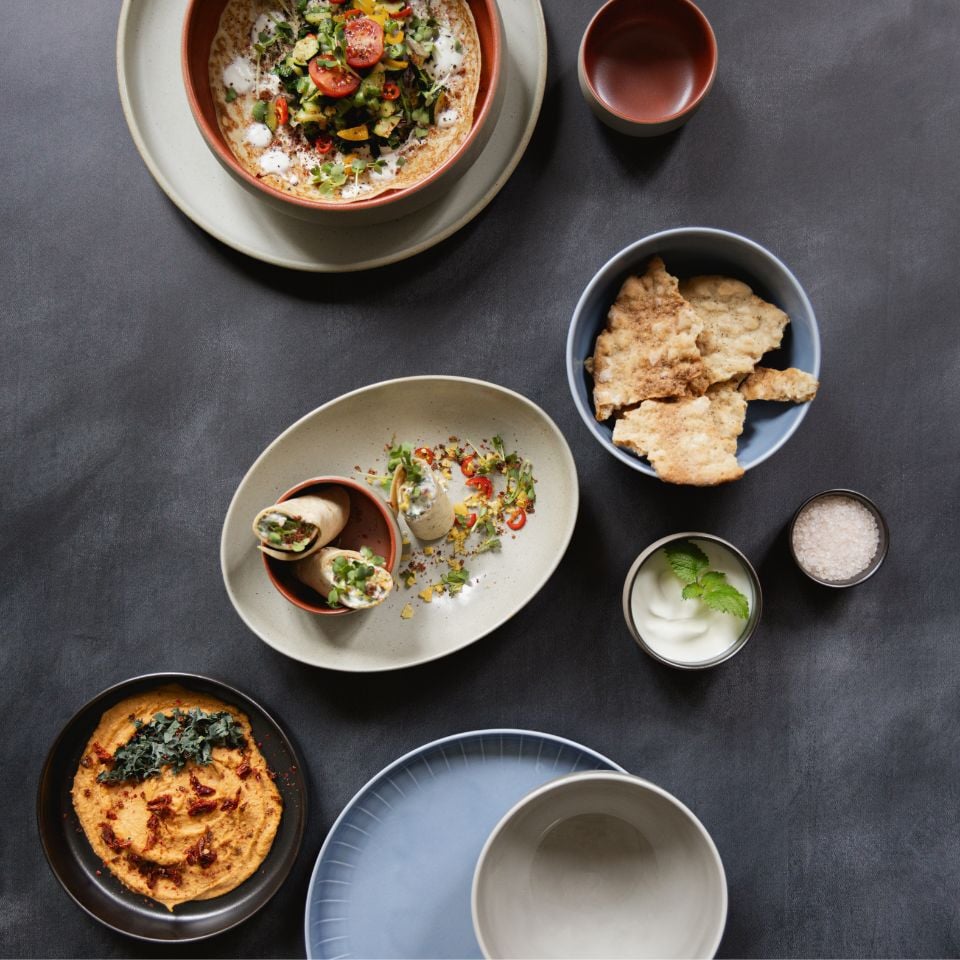 An overhead view of a variety of dishes in stoneware bowls and plates, featuring hummus, flatbread, stuffed wraps, dips, and salad, styled on a dark matte table surface.