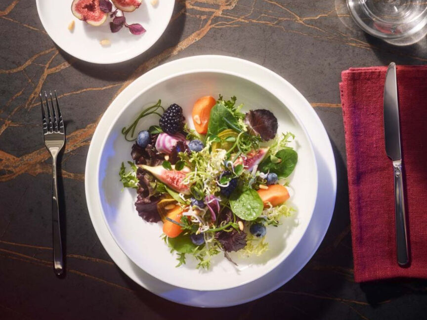 Weiße Schüssel mit buntem Salat platziert auf großem weißen Teller mit silbernem Besteck