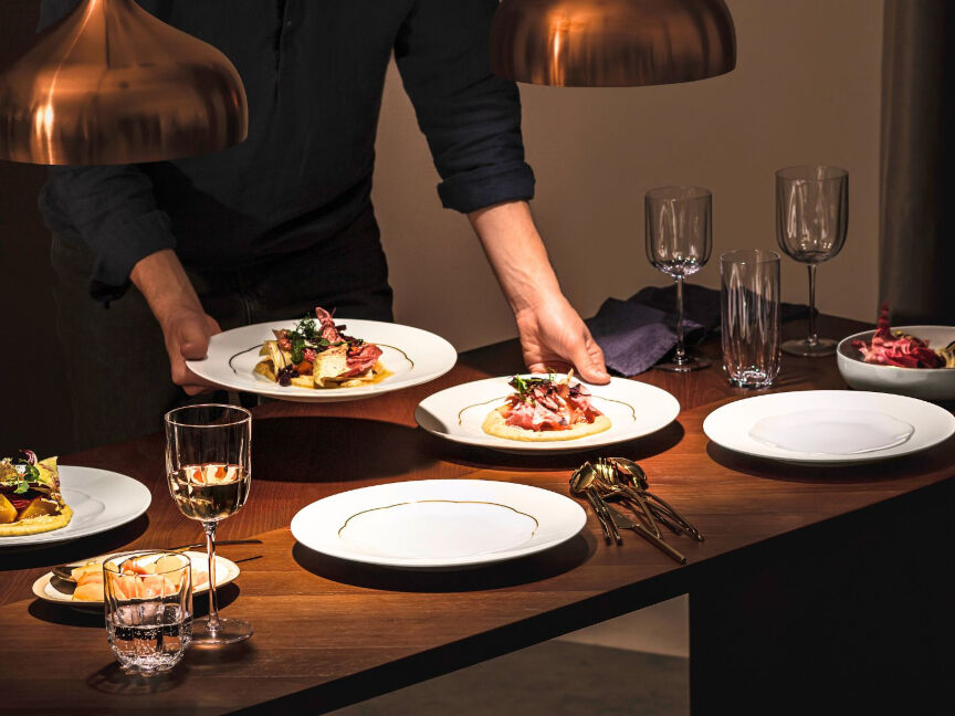 Festlich gedeckter Holztisch mit Tellerset in weiß mit Goldrand der Sonetto-Kollektion und goldenem Besteck