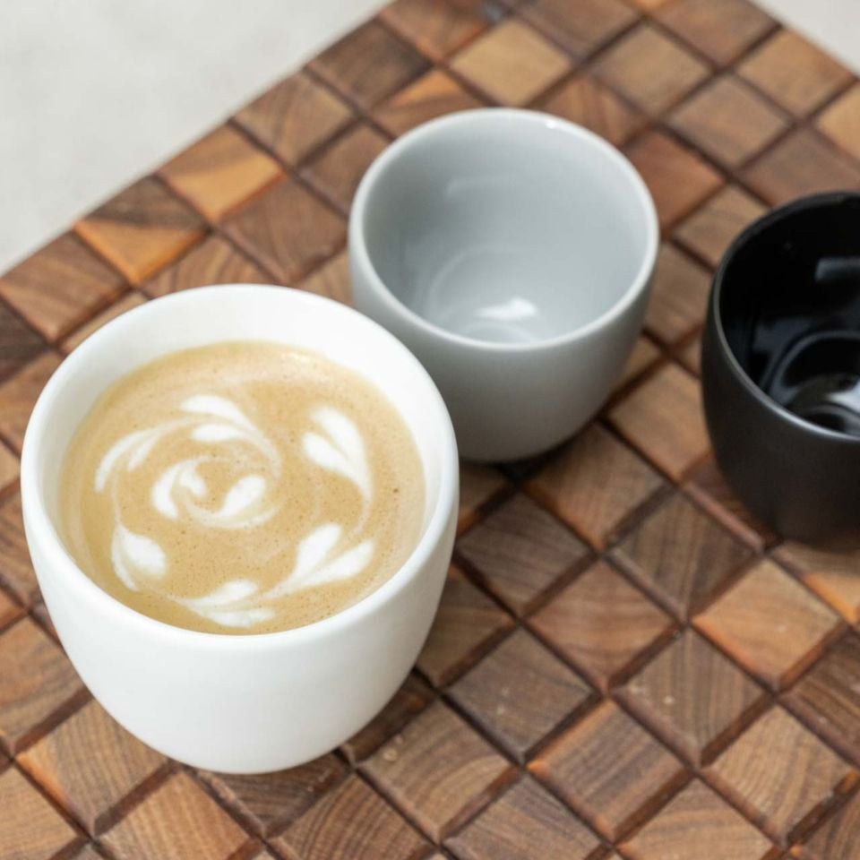Close-up of three small ceramic cups in white, grey, and black, with one filled with cappuccino and latte art, on a patterned wooden tray.