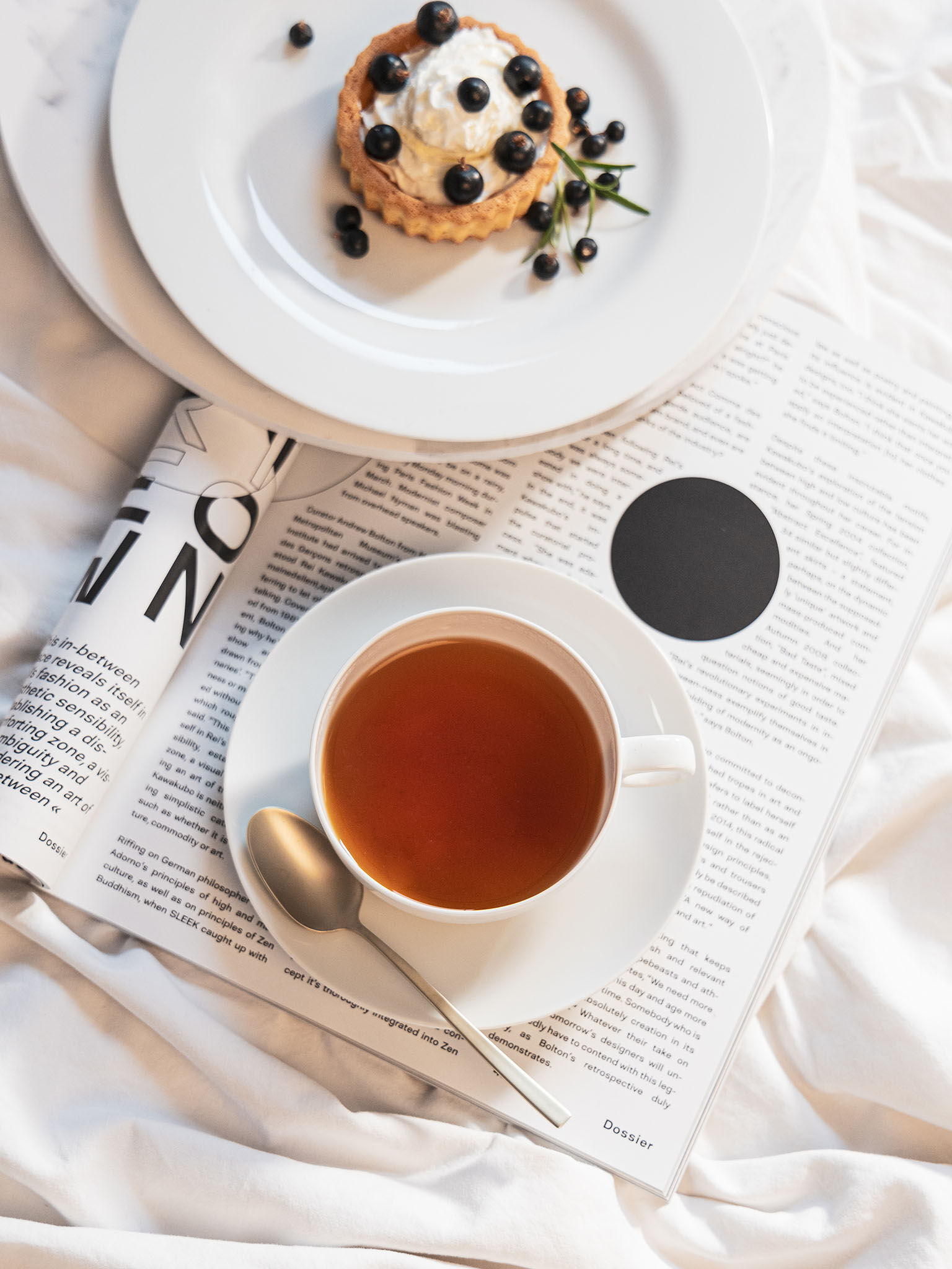 Moon teacup with saucer filled with tea and breakfast plate with small sponge cake placed in bed with a magazine.
