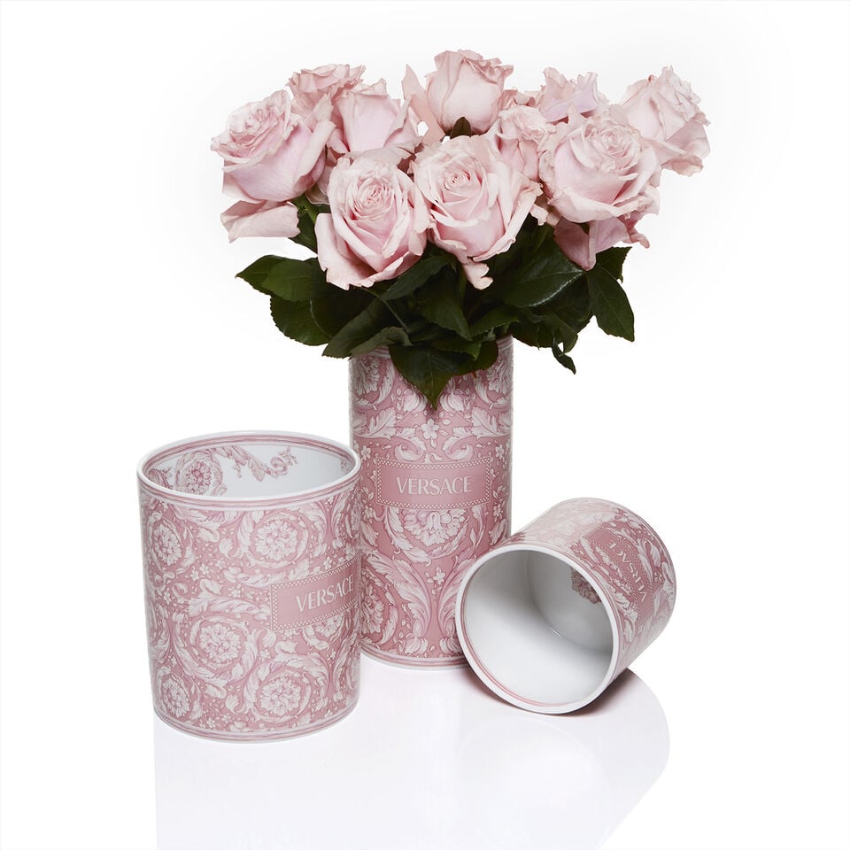 Set of three cylindrical Versace porcelain vases in pastel pink with intricate baroque floral patterns and the Versace logo, one of them filled with a bouquet of pale pink roses.