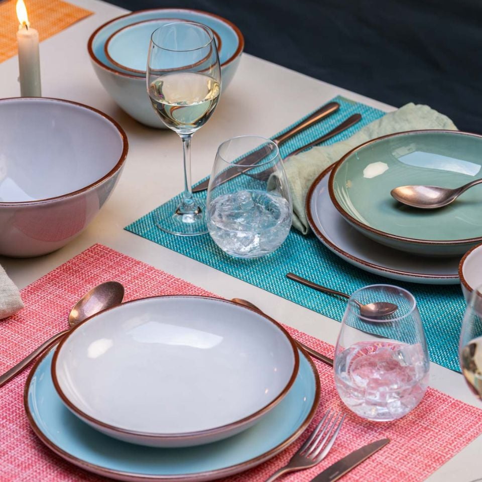 A colorful outdoor table with plates in soft hues, pink and blue placemats, and wine glasses with water and white wine.