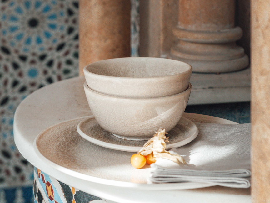 A beige ceramic bowl by Rosenthal with a matching plate on a round side table, with oriental mosaic tiles and a stone column in the background.