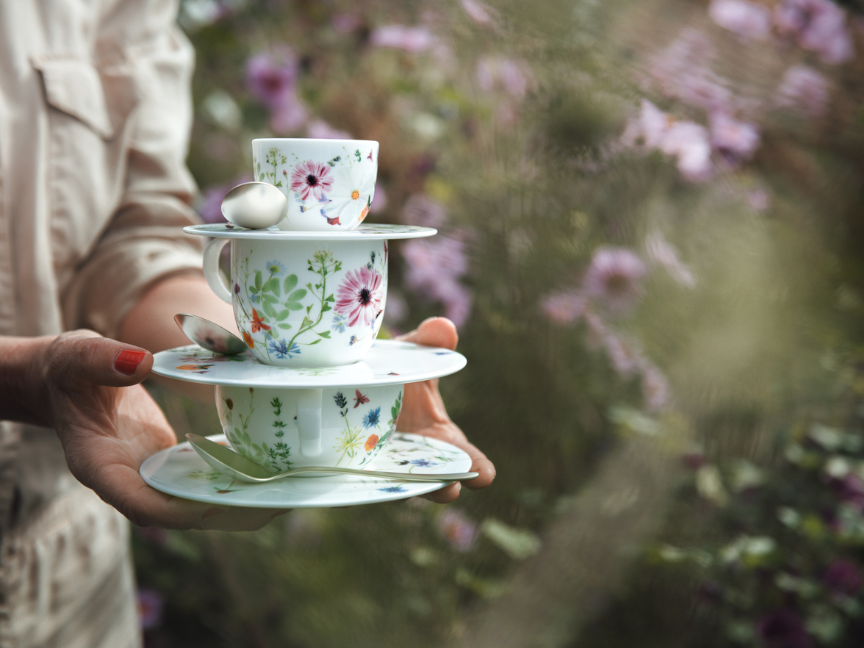 Kaffeetassenservice mit Blumendekor wird von Frau durch Blumenwiese getragen