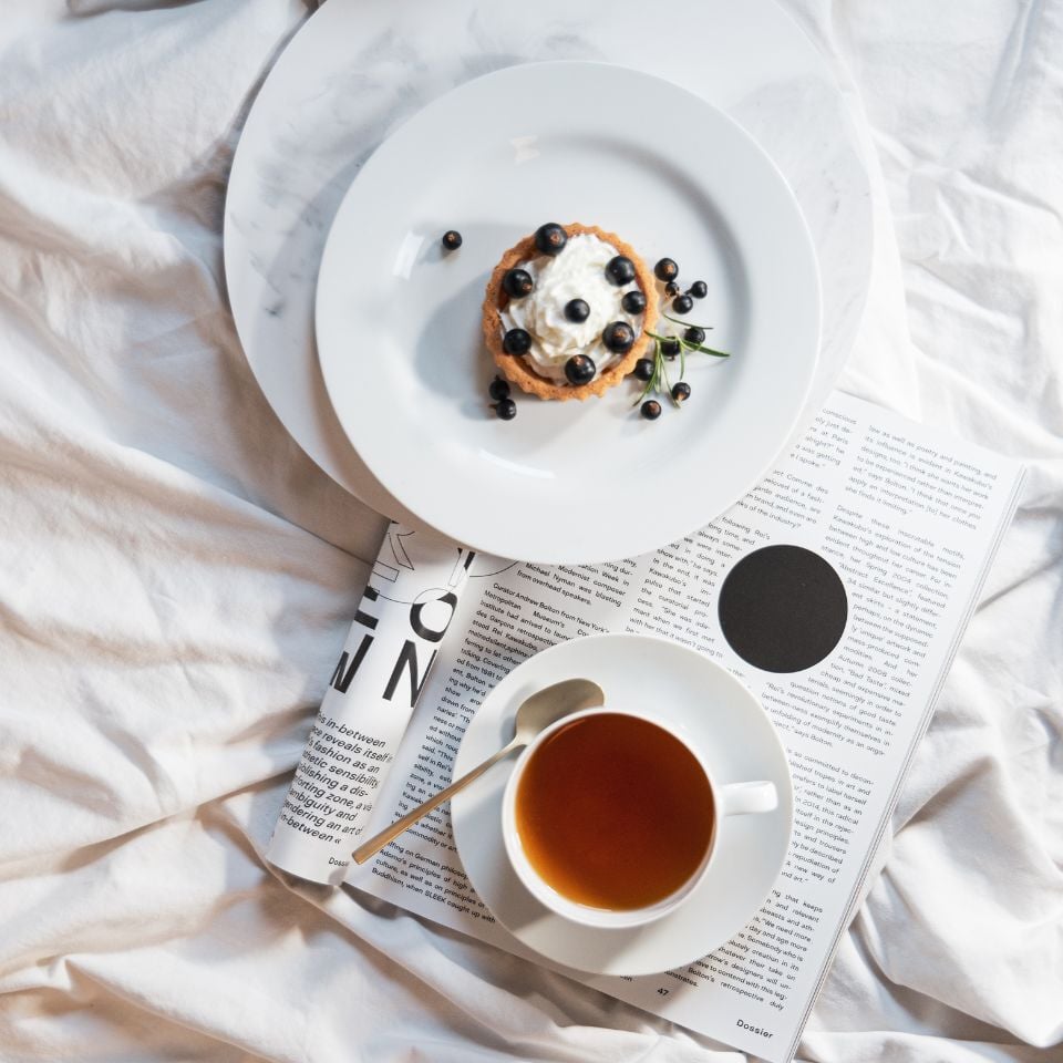 A white plate with a berry tart and a cup of tea on a bed, placed over an open magazine in soft daylight.