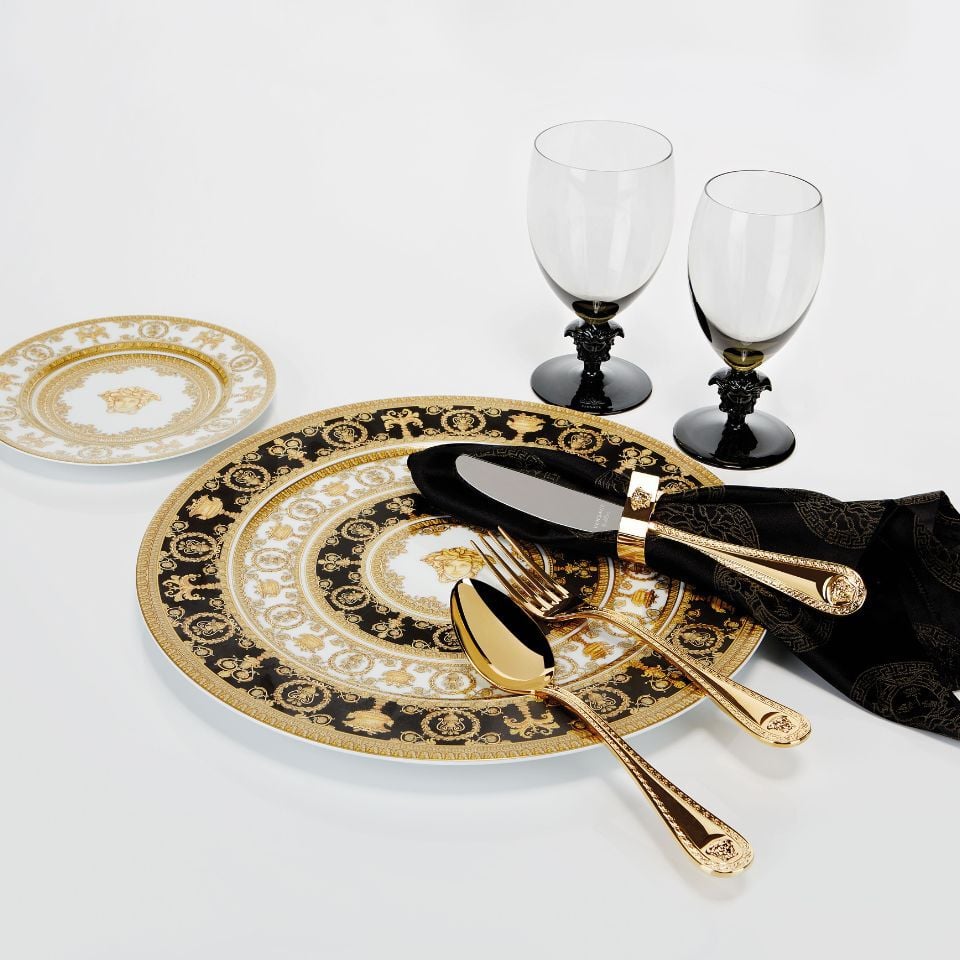 Versace's I Love Baroque dinnerware collection with lavish black and gold design, Medusa motifs, and matching gold cutlery on a crisp white background.