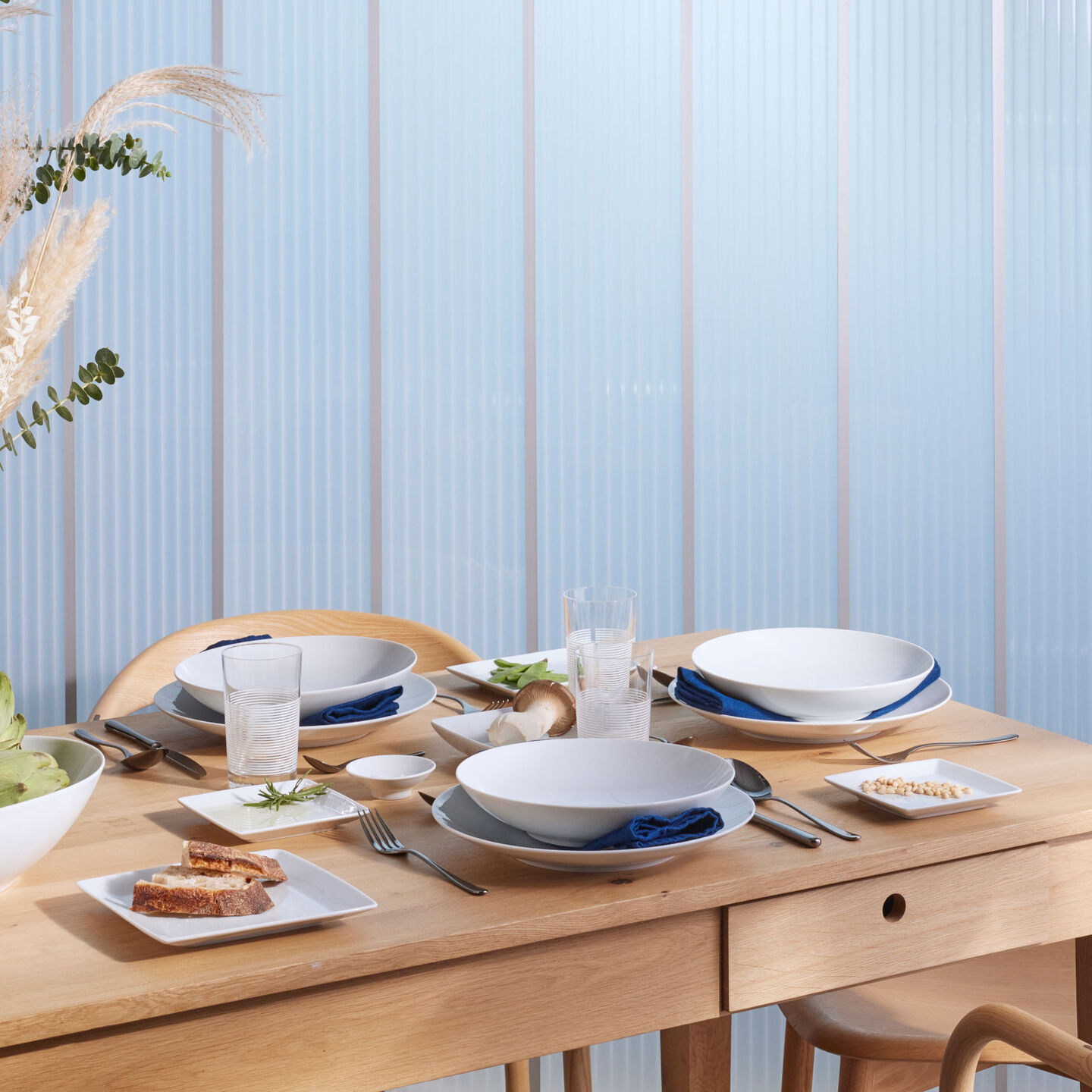 A wooden table set with Thomas Loft White deep plates, flat plates & blue napkins