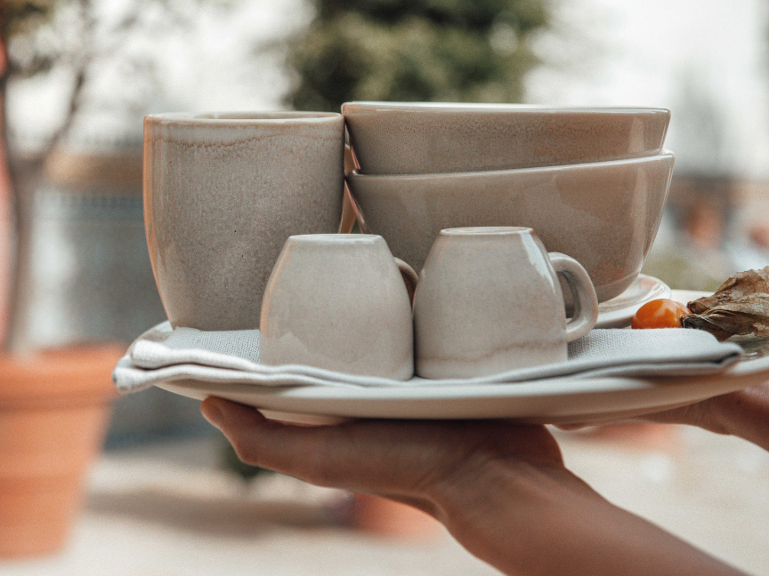 H&auml;nde tragen einen Stapel Rosenthal Steinzeug-Geschirr mit Tellern, Schalen, Becher und zwei Espressotassen vor unscharfem Outdoor-Hintergrund.