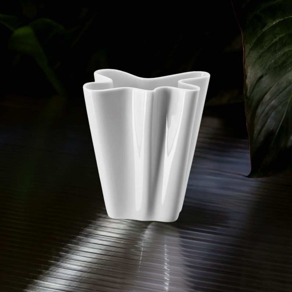 A white sculptural vase with softly folded contours, resting on a metallic surface.