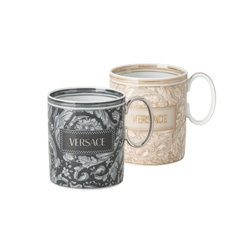 Pair of Versace porcelain mugs featuring detailed baroque leaf patterns, one in black and silver, the other in beige and gold, both with the Versace logo prominently displayed.