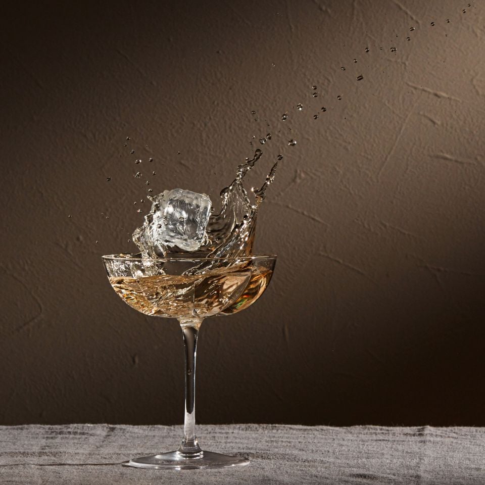 A coupe glass with golden liquid splashing over the rim as an ice cube drops in midair.
