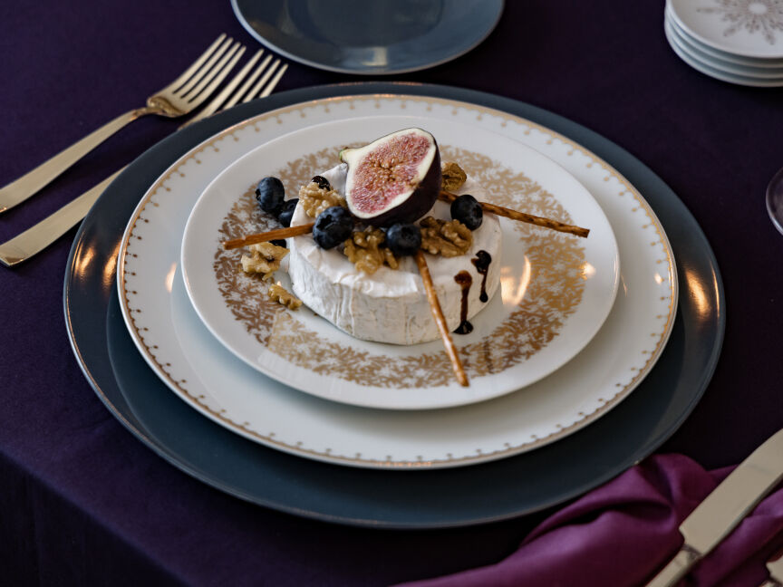 Kleiner Speiseteller platziert auf großem Speiseteller der TAC Festive Celebration Kollektion in weiß mit goldenen Dekoren. Auf dem Teller ist ein Camembert mit Wallnüssen, Feige und Blaubeeren.