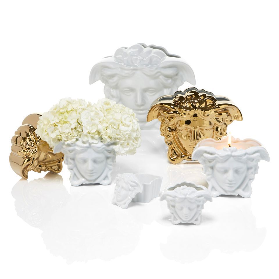 Collection of decorative Versace Medusa head accessories in white and gold finishes, including vases, candle holders, and a flower pot filled with white hydrangeas.