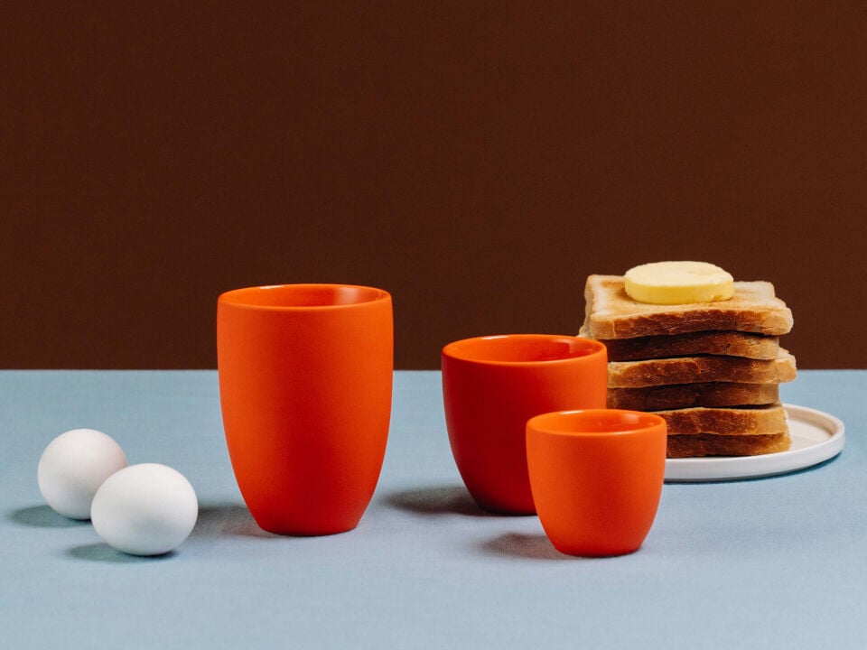 Three orange cups in different sizes with eggs and toasts aside on blue-coloured table cloth and brown background